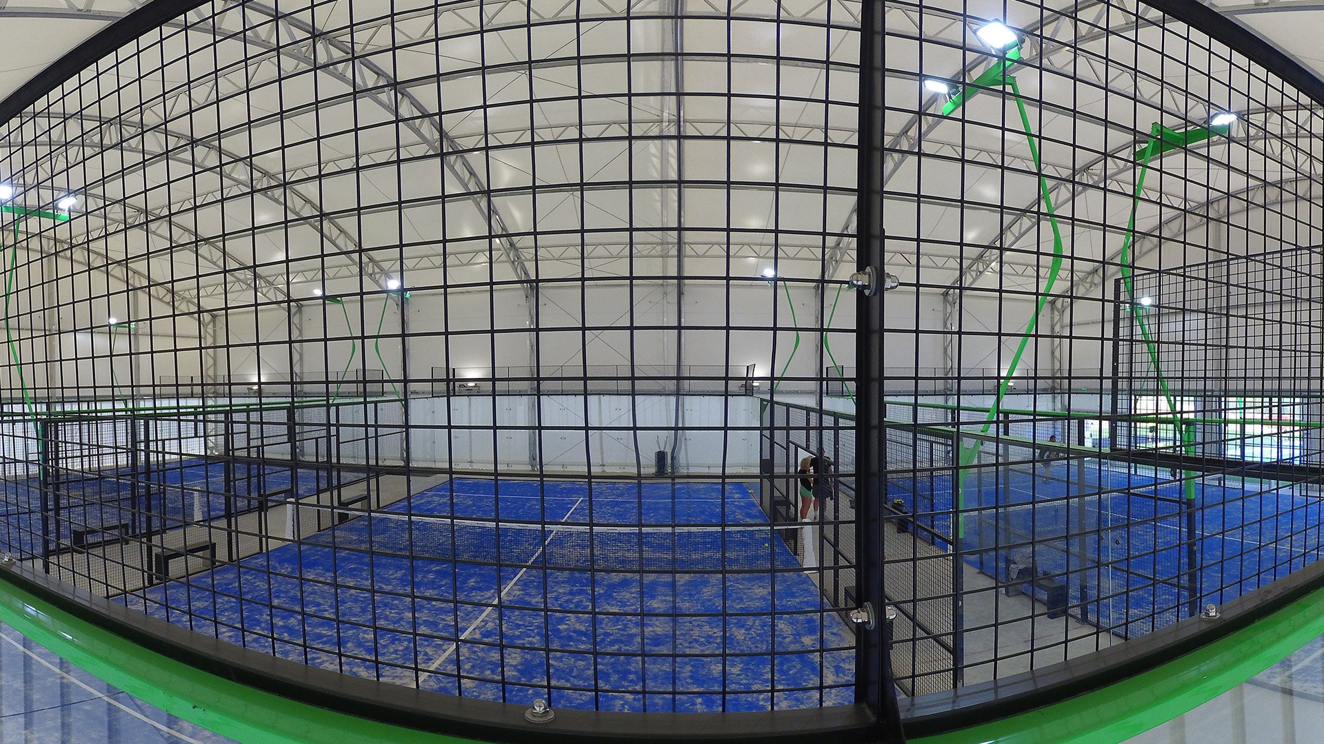 Padel court interior with sand-covered blue playing surface, enclosed by black mesh fencing and illuminated by overhead lights, showcasing the unique design for enhanced gameplay.