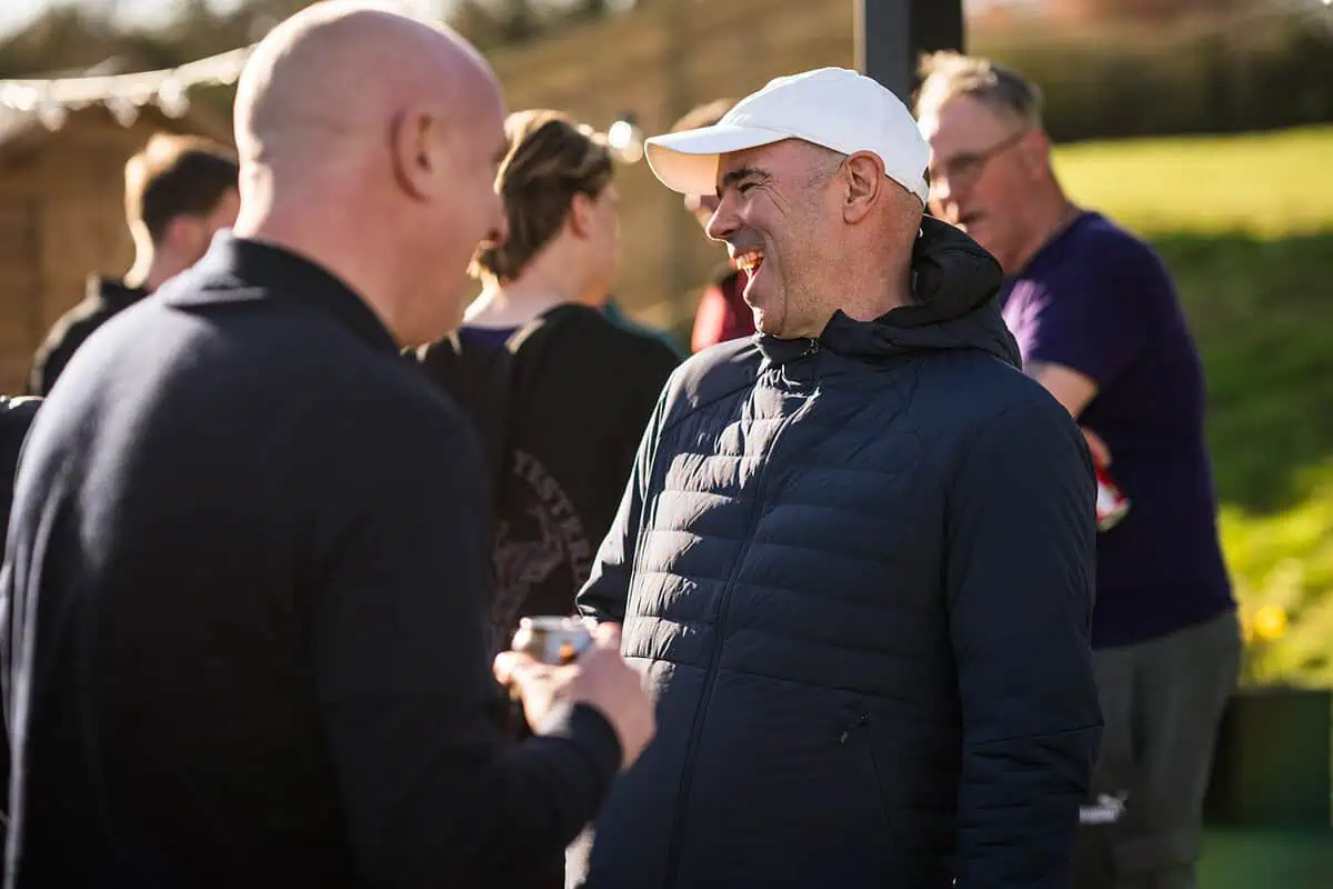 Two men engaging in conversation outdoors, one wearing a white cap and a dark jacket, with a group of people socialising in the background, illustrating a lively padel court environment in Kent.