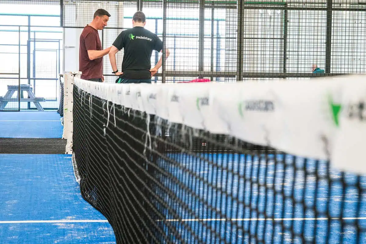 Two men discussing padel court construction beside a net in a blue indoor padel court environment, featuring visible branding on the net and surrounding infrastructure.