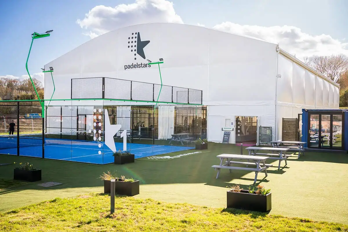 Padel court installation at Padelstars facility in Bristol, featuring a white structure, glass panels, and blue playing surface, surrounded by landscaped areas and seating.